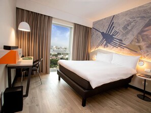 A hotel room with a large bed, a desk, and a view of a cityscape through a window.
