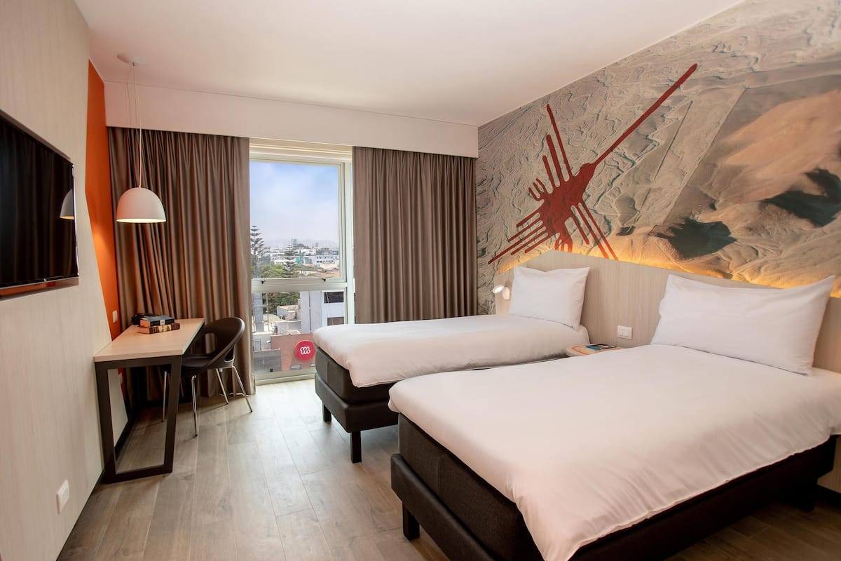 Une chambre d’hôtel avec deux lits, un bureau et une grande fenêtre avec des rideaux.