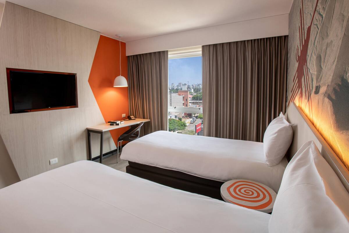 Een hotelkamer met twee bedden, een bureau met een stoel, een aan de muur bevestigde televisie en een groot raam met gordijnen.