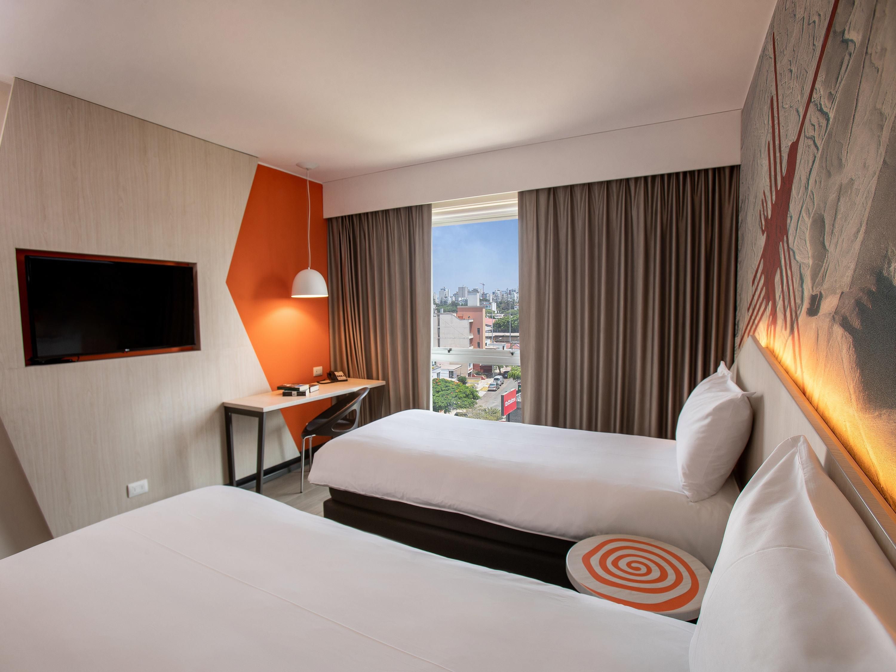 A hotel room with two beds, a desk with a chair, a TV mounted on the wall, and a large window with curtains.