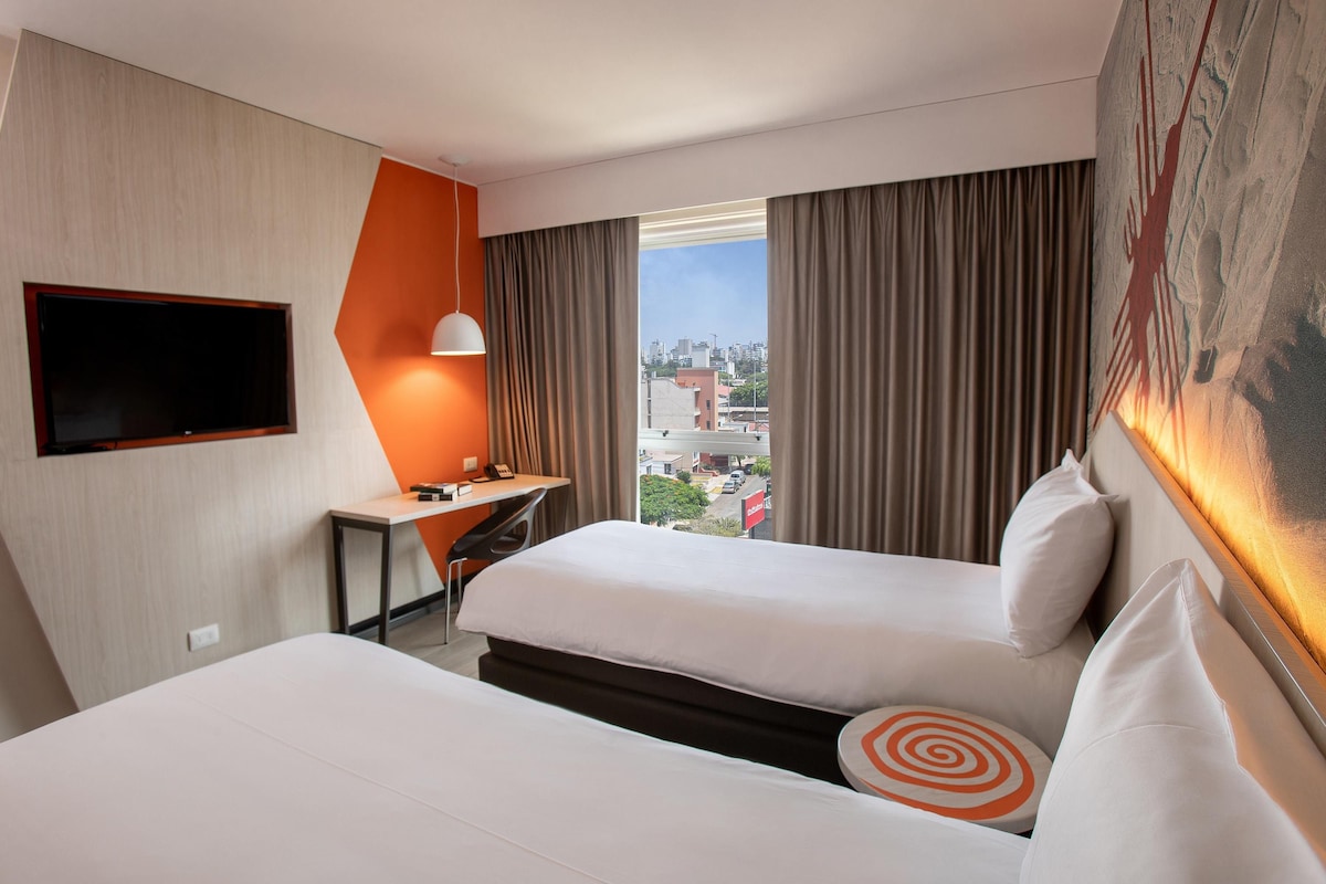 Une chambre d’hôtel avec deux lits, un bureau avec une chaise, un téléviseur fixé au mur et une grande fenêtre avec des rideaux.