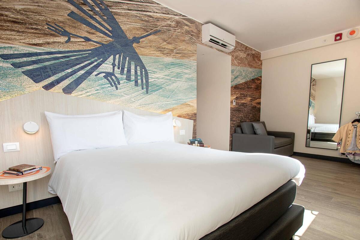 Une chambre d’hôtel avec un lit, une fresque murale, un canapé gris, une petite table ronde et un miroir.