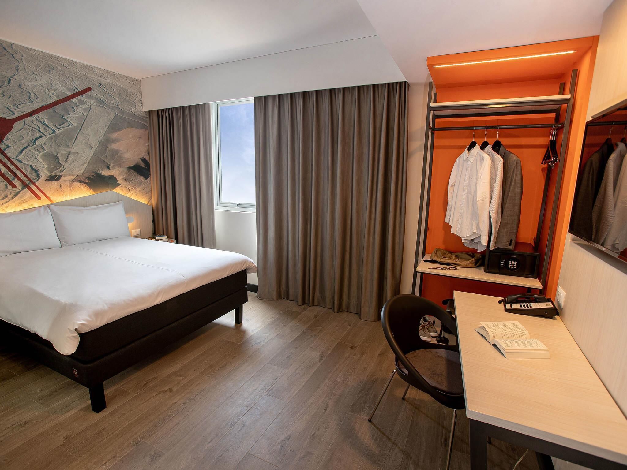 A hotel room with a bed, a desk with a chair, a coat rack with clothes, a window with curtains, and a wall with a painting.