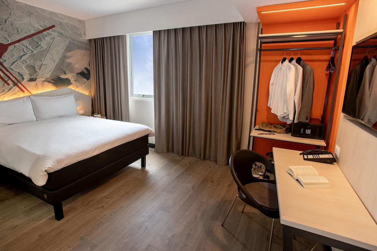 A hotel room with a bed, a desk with a chair, a coat rack with clothes, a window with curtains, and a wall with a painting.