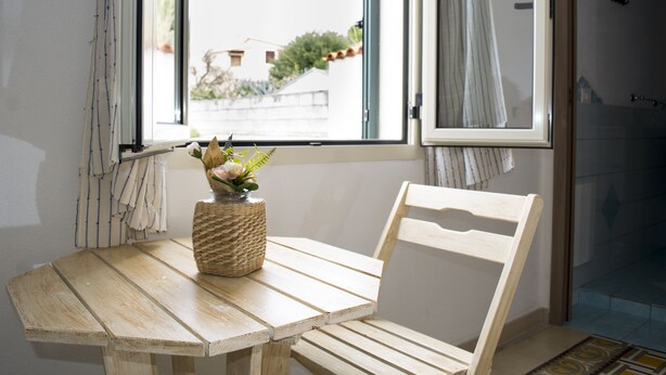 A wooden table with a woven basket containing a plant and flowers, a wooden chair, and a window with curtains. - Bed And Breakfast Villa Bianca (Orosei)
