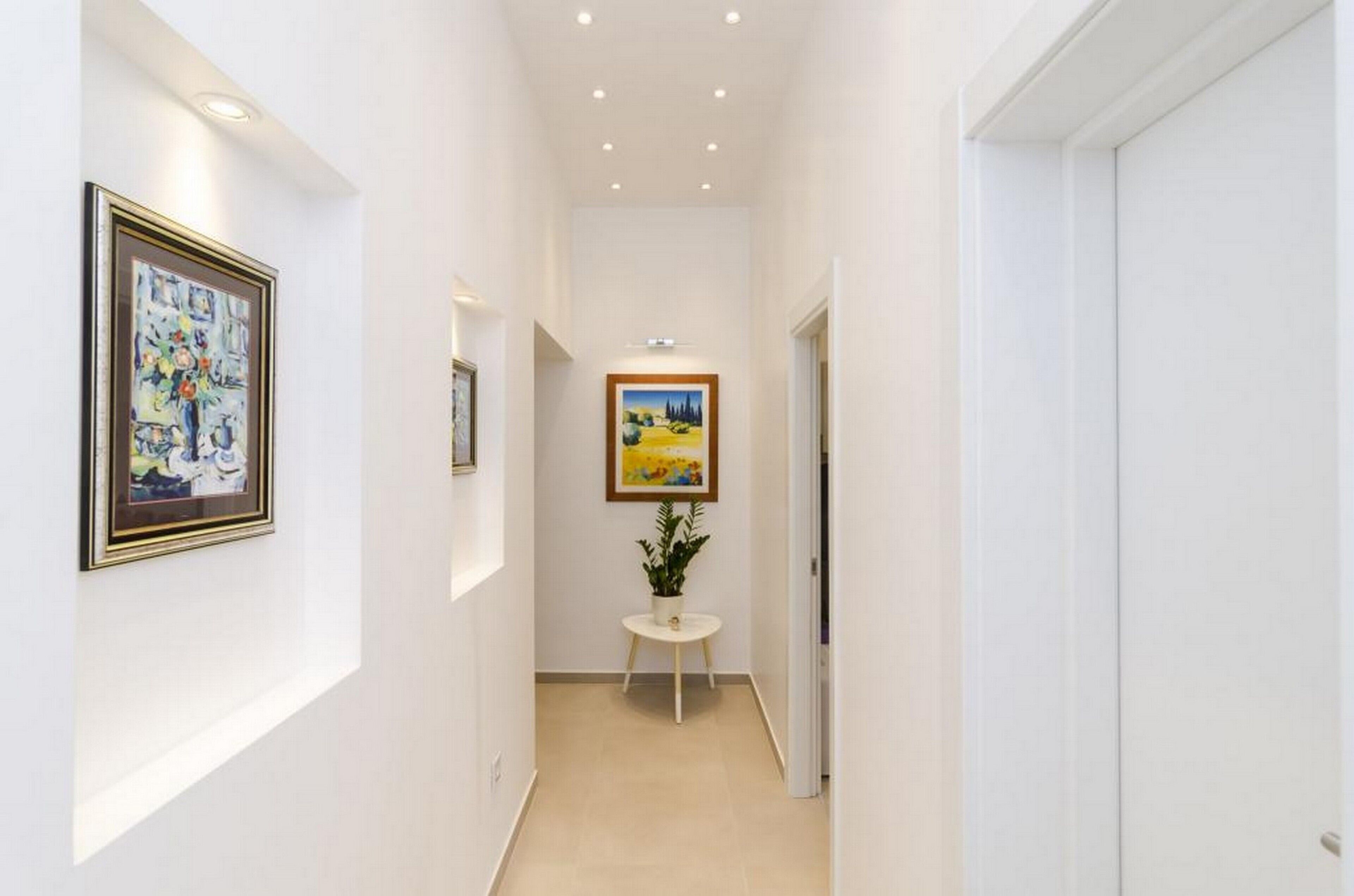 A narrow hallway with white walls, two framed paintings, a small round table with a plant, and a door at the end.