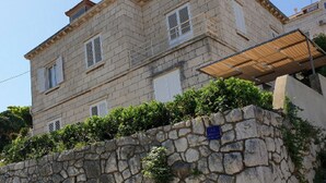 Ein mehrstöckiges Steinhaus mit einer Steinmauer im Vordergrund und einer überdachten Außenfläche unter blauem Himmel.