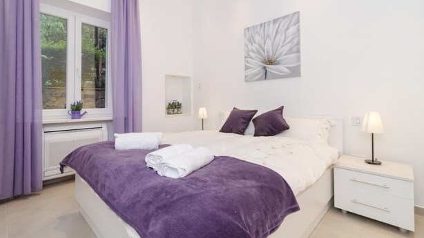 Ein Schlafzimmer mit einem Bett, weißen Nachttischen, einem Fenster mit violetten Vorhängen, einem Blumenbild an der Wand und einer Heizung.
