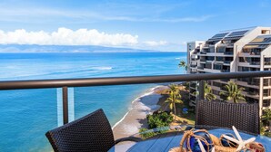 A balcony with a view of the ocean and a beach, featuring a dining table with a woven chair and a decorative bowl. - Stunning, Oceanfront Deluxe Penthouse with Island Views - Sands of Kahana 282 (Lahaina)