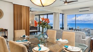 A dining area with a glass table, chairs, and a vase of flowers. There is a sofa and a view of the ocean in the background. - Stunning, Oceanfront Deluxe Penthouse with Island Views - Sands of Kahana 282 (Lahaina)