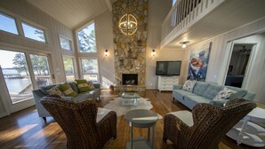 A spacious living room with a stone fireplace, wicker furniture, and large windows overlooking a deck. - 6000 SF Lakehouse with Hot Tub Games Dock and Ramp (Camden)