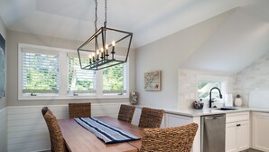 Un comedor con mesa de madera y sillas de mimbre, una lámpara moderna y una cocina con armarios blancos y un lavavajillas de acero inoxidable.