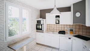 Ett modernt kök med vita skåp, en inbyggd mikrovågsugn, en ugn och en diskho. Ett fönster släpper in naturligt ljus och ger utsikt över grönområden utanför.