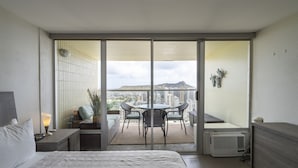 A hotel room with a bed, a bedside table with a lamp, a small table with chairs, and a view of the city through large sliding doors.