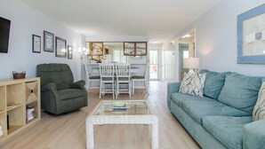 Ein Wohnzimmer mit einem grünen Sessel, einem blauen Sofa, einem Glastisch und einem Essbereich mit weißem Tisch und Stühlen.