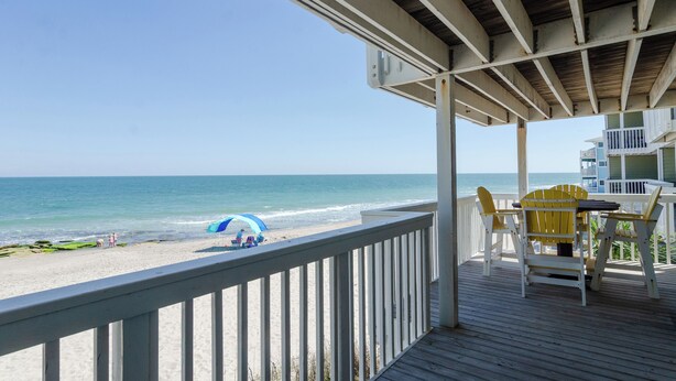 Eine Holzterrasse mit Meerblick, ein Tisch mit Stühlen und ein blauer Sonnenschirm am Strand.