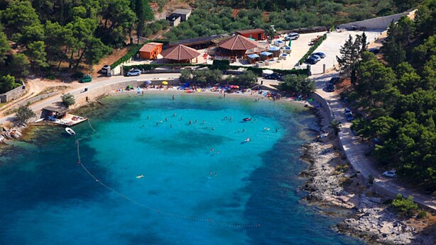 Vue aérienne d’une zone côtière avec une plage, plusieurs bâtiments et une mer bleue et limpide.