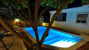 Une piscine avec un arbre au premier plan, un bâtiment blanc doté de fenêtres et des meubles d’extérieur.