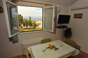 A room with a table set for two, a television, and a window with a view of buildings and trees. - Villa Anni (Sutivan)