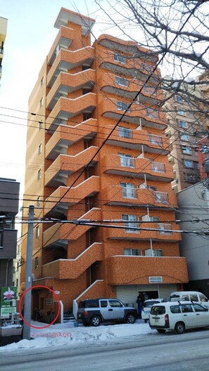 Un immeuble résidentiel à plusieurs étages, doté de balcons, une aire de stationnement avec des voitures et une rue avec un panneau indicateur.