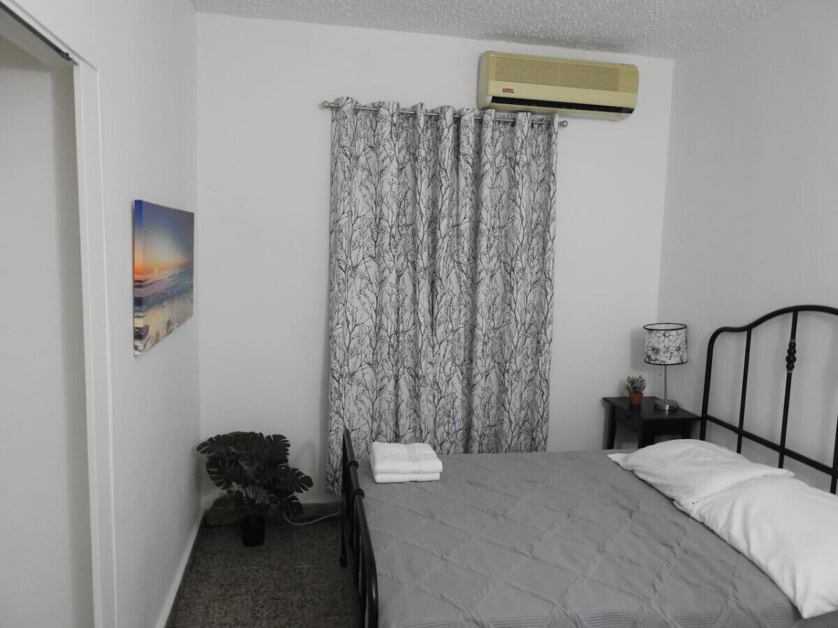 Un dormitorio con cama, mesita de noche, planta en maceta, aire acondicionado de pared y un cuadro en la pared.
