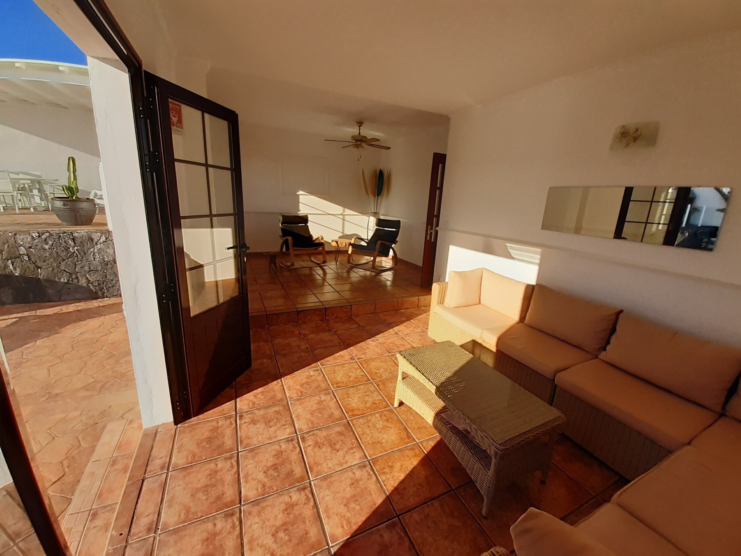An open-plan living area with a sofa, wicker table, and chairs. A ceiling fan is visible, and there is a potted plant in the background.