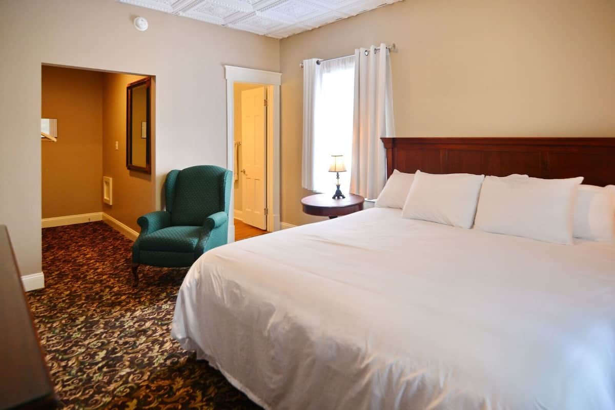 Une chambre d’hôtel avec un grand lit, un fauteuil vert, une petite table avec une lampe et une porte donnant sur une autre pièce.