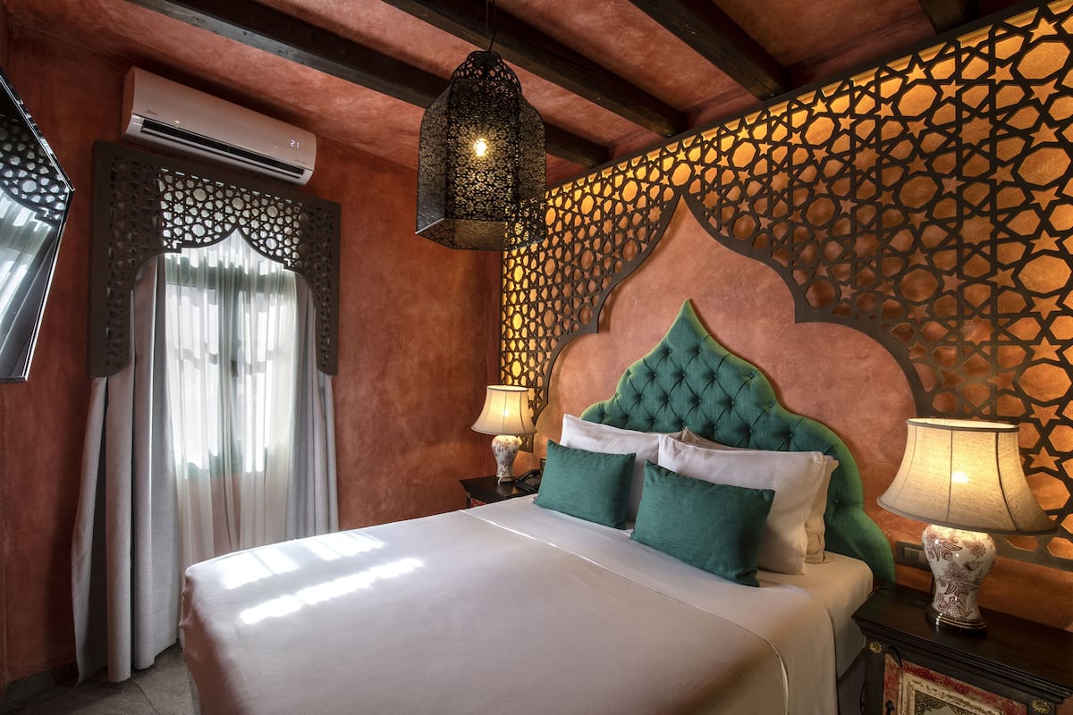 Een net opgemaakt bed met groene en witte kussens, een decoratieve wand met geometrische patronen en een hanglamp.