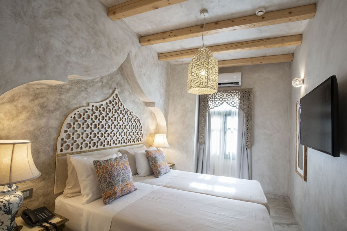 Een slaapkamer met twee bedden, een decoratief hoofdbord, een hanglamp, een telefoon op het nachtkastje en een tv aan de muur.