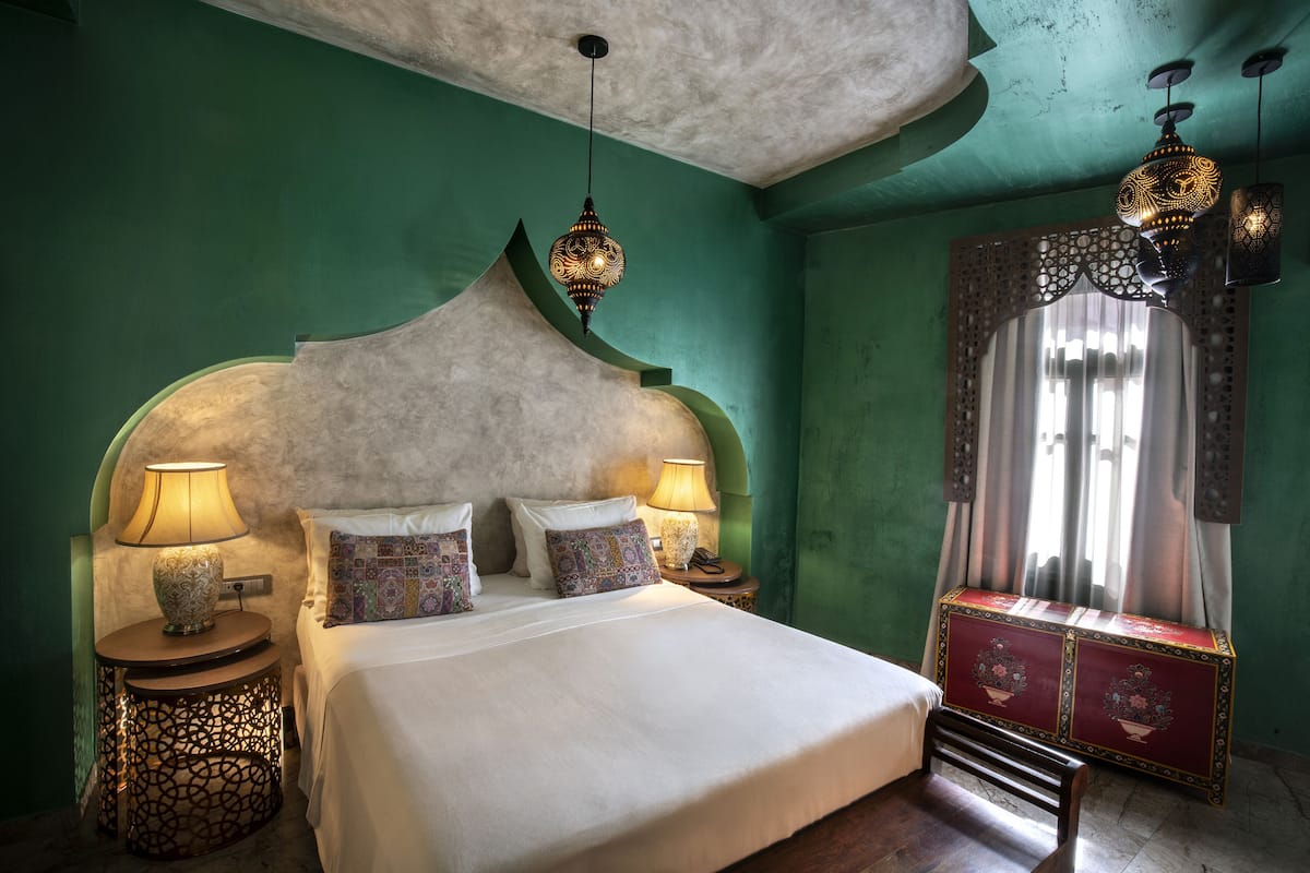 Een net opgemaakt bed met gekleurde kussens, een nachtkastje met een lamp en een decoratieve kast.