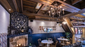 Un restaurante acogedor con chimenea de piedra, vigas de madera en el techo y un candelabro. Mesas y sillas dispuestas para comer.
