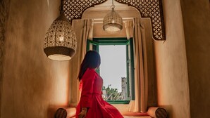 Una mujer con un vestido rojo parada junto a una ventana con cortinas estampadas, una lámpara colgante tejida y un alféizar de ventana con marco verde.