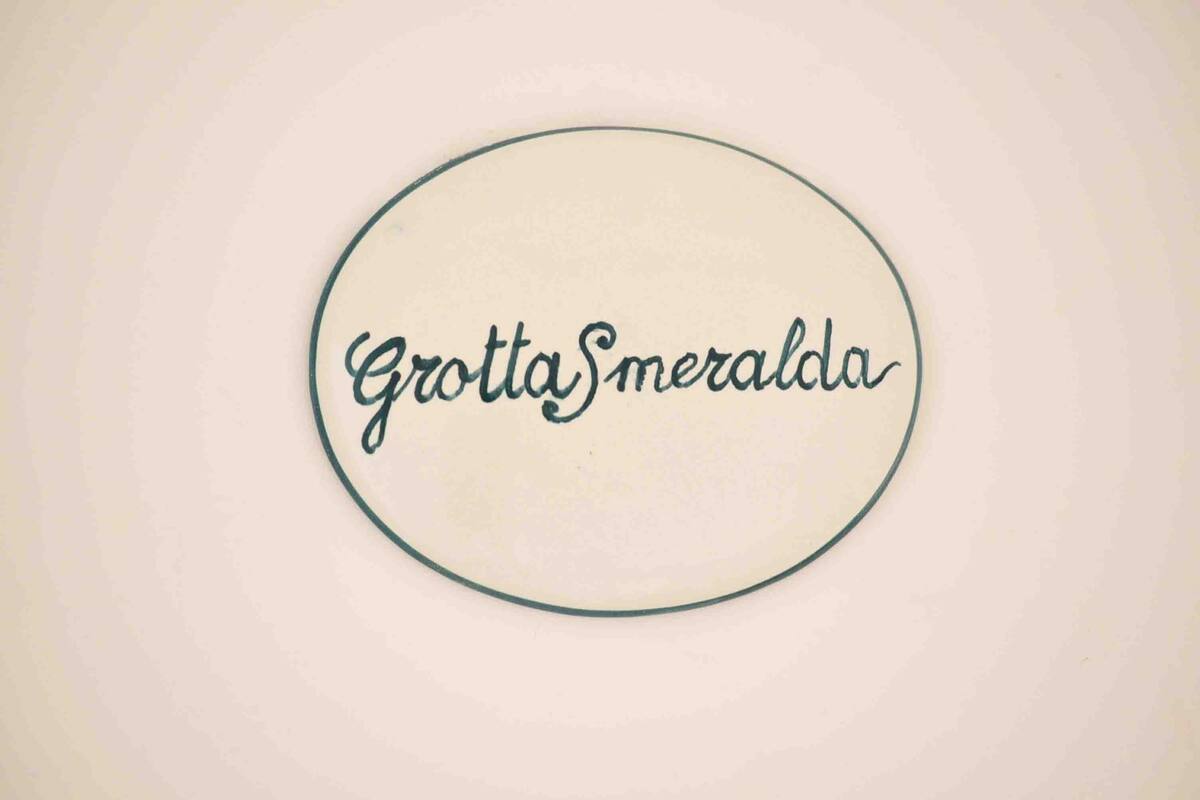 Un panneau circulaire sur lequel on peut lire le texte « GrottaSmeralda » et qui est placé sur un fond uni.