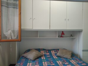 Une chambre à coucher avec un lit, deux oreillers, une couvre-lit à motifs, une lampe rouge posée sur une étagère et une armoire blanche.