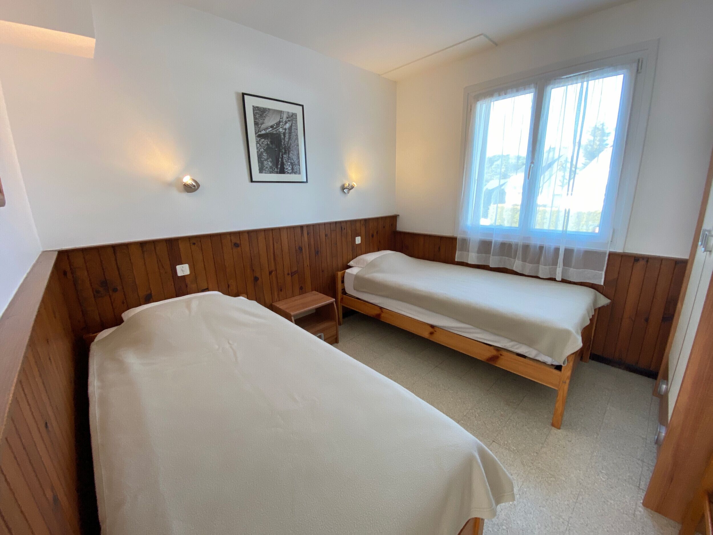 A room with two single beds, a small table, a window with sheer curtains, and a framed picture on the wall.