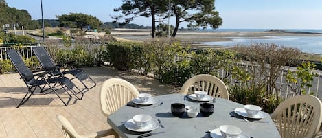 Een terras met een tafel gedekt voor vier personen, omringd door stoelen, met uitzicht op een strand en heuvels in de verte.