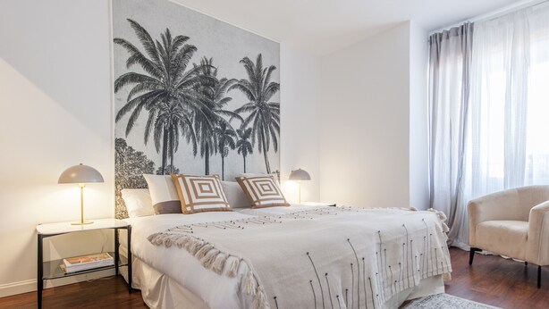 Een slaapkamer met een groot bed, twee nachtkastjes met lampen, een stoel, een groot wanddecoratie van palmbomen en een raam met gordijnen.