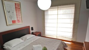 A neatly made bed with a striped bedspread and a white pillow, a bedside table with a lamp, and a large window with blinds. - Beach House - Between Porto, Braga and Viana do Castelo (Esposende)