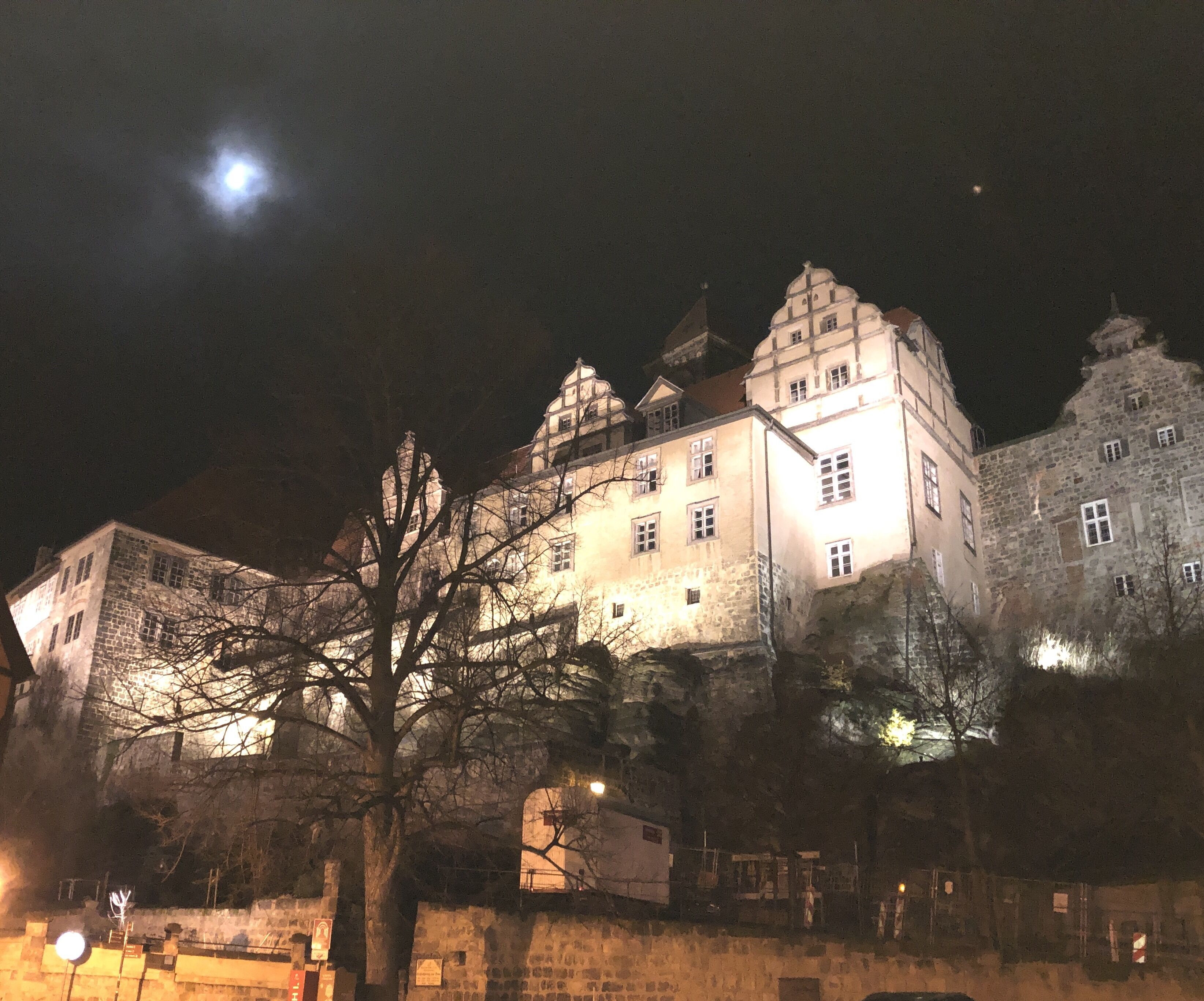 A historic building with multiple towers and a prominent central tower, surrounded by stone walls and a tree in the foreground.