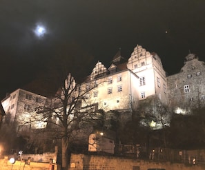 Um edifício histórico com várias torres, incluindo uma torre central imponente, rodeado por muralhas de pedra e uma árvore em primeiro plano.