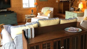 L’intérieur d’un chalet accueillant, avec un plafond en bois, un téléviseur, un canapé, un fauteuil, une petite table et une lampe.