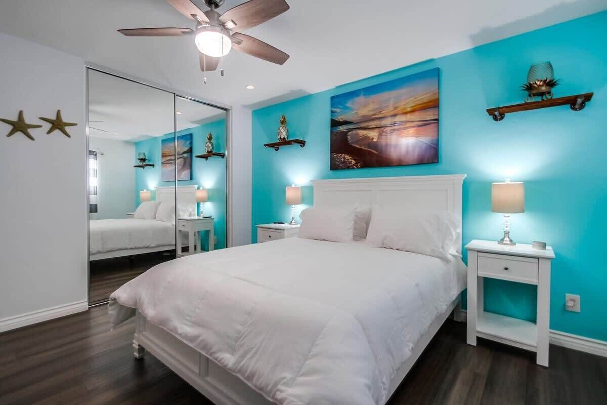 Ein Schlafzimmer mit einem großen Bett, einem Deckenventilator, einem Spiegel, einem Nachttisch mit Lampe und einem Wandbild.