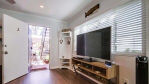 Een kamer met een houten vloer, een grote flatscreen televisie op een houten voetstuk, een deur met het nummer 2 en een raam met jaloezieën.