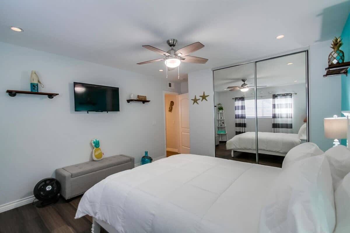 A bedroom with a bed, a ceiling fan, a TV, a mirror, and a window with blinds.