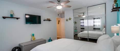 A bedroom with a bed, a ceiling fan, a TV, a mirror, and a window with blinds.