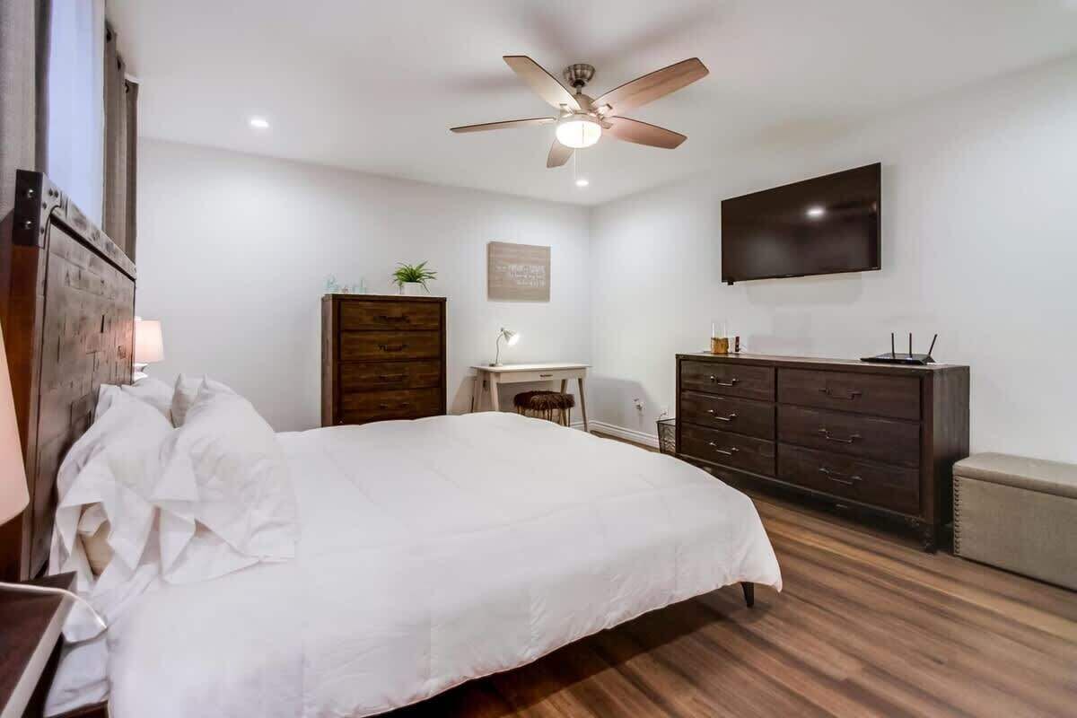 A bedroom with a bed, a desk, a dresser, a ceiling fan, and a TV mounted on the wall.