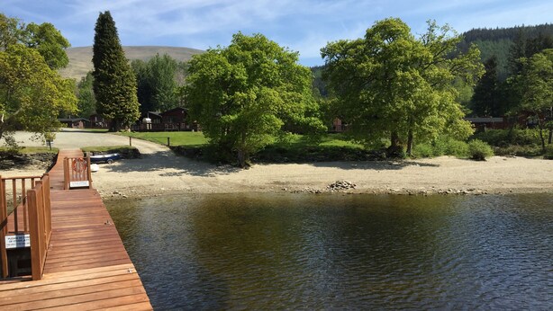 A wooden dock extending over a calm river, surrounded by lush greenery and a distant hill. - Lomond Lodge 20, Luxury Log cabin on the banks of Loch Lomond (Loch Lomond)
