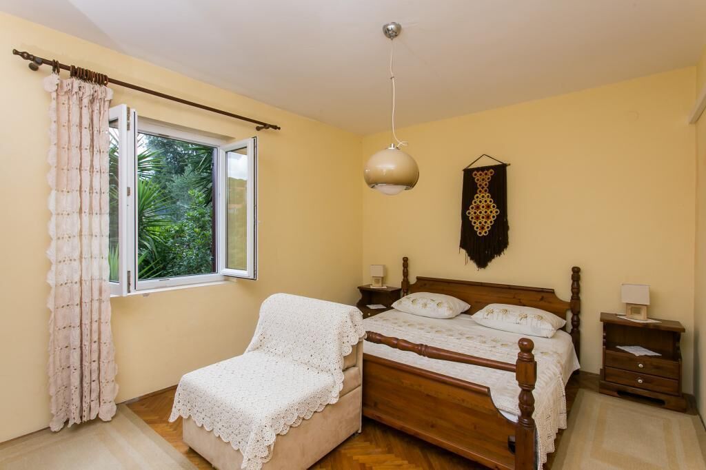 A bedroom with a wooden bed, a small bedside table, a nightstand, a window with curtains, and a hanging wall decoration.