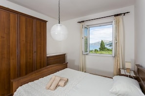 A bedroom with a large wooden wardrobe, a bedside table with a lamp, a bed with a white bedspread and rolled towels, and a window with a view of a lake and mountains.