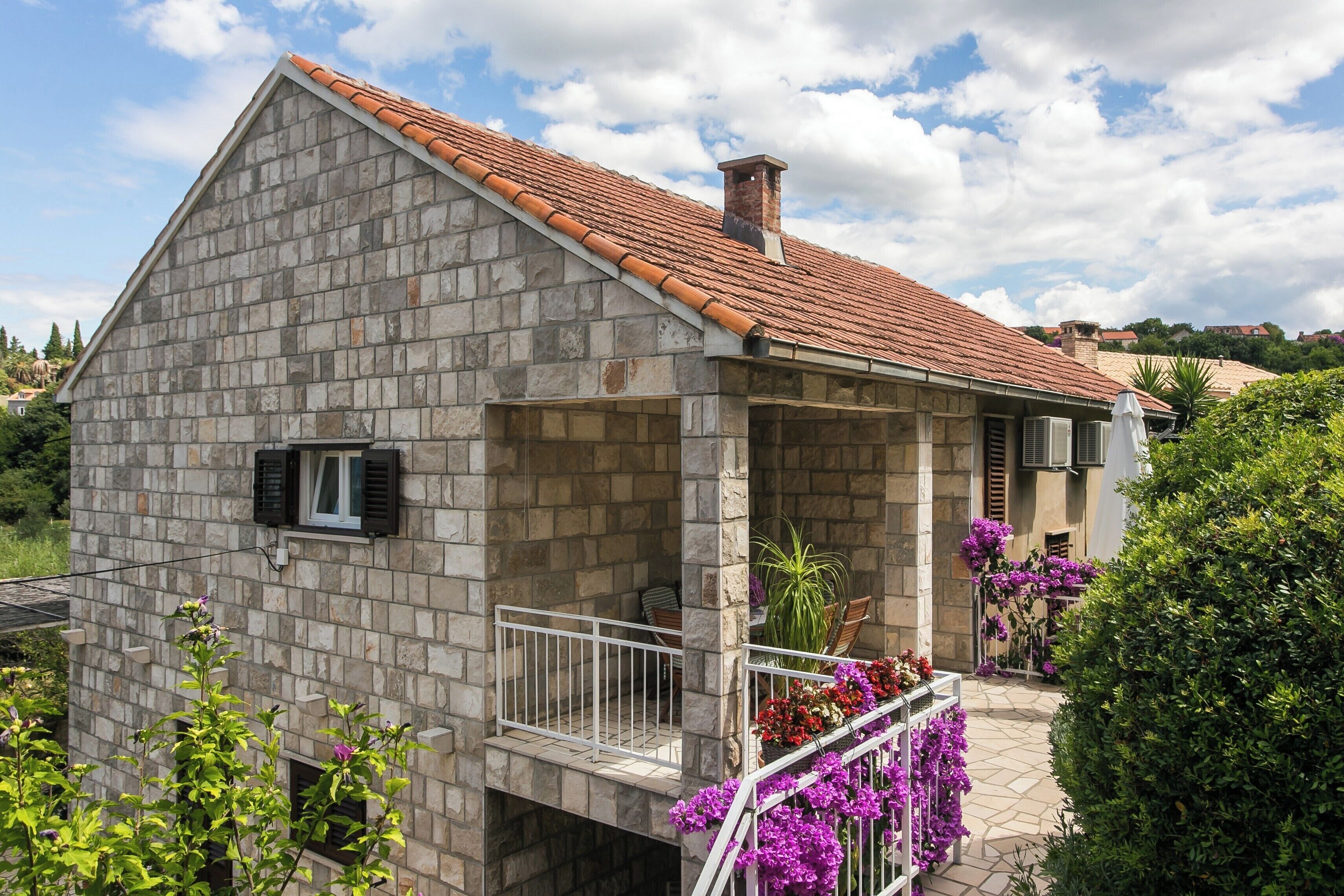 A stone house with a tiled roof, a balcony with a railing, and a small garden area with flowers.